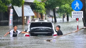 Ô tô ngập sâu giữa đường phố khi mưa lớn, trong lúc hai người tìm cách hỗ trợ đưa phương tiện ra khỏi vùng nước.