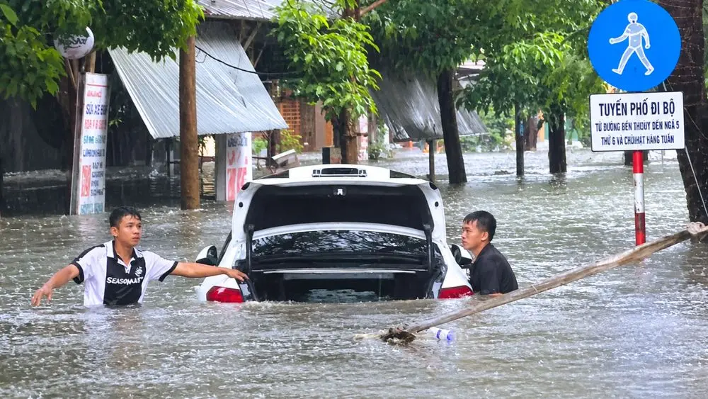 Ô tô ngập sâu giữa đường phố khi mưa lớn, trong lúc hai người tìm cách hỗ trợ đưa phương tiện ra khỏi vùng nước.